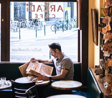 Un homme est assis à une table dans un café, lisant un journal, avec une vue sur une rue animée et des vélos à l'extérieur.