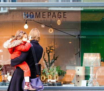 Une mère avec un enfant en manteau rouge regarde une vitrine de magasin de meubles portant le nom 'HOMEPAGE'.
