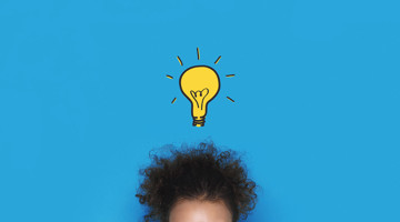 Een persoon met donker, krullend haar kijkt omhoog naar een brandende gloeilamp boven haar, tegen een heldere blauwe achtergrond.