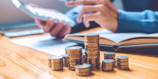Une main avec des pièces de monnaie et une calculatrice, entourée de piles de pièces et d'un livre ouvert sur une table en bois.